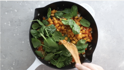 A hand stirs fresh spinach, diced sweet potatoes, and bell peppers in a white-handled black skillet on a light gray countertop, preparing savory Breakfast Meal Prep Bowls.