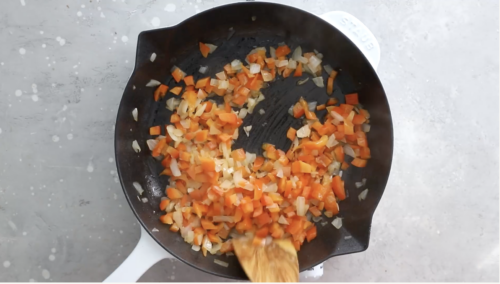 Chopped orange bell peppers and white onions are being sautéed in a cast iron skillet with a wooden spoon, perfect for adding vibrant flavor to your Breakfast Meal Prep Bowls.