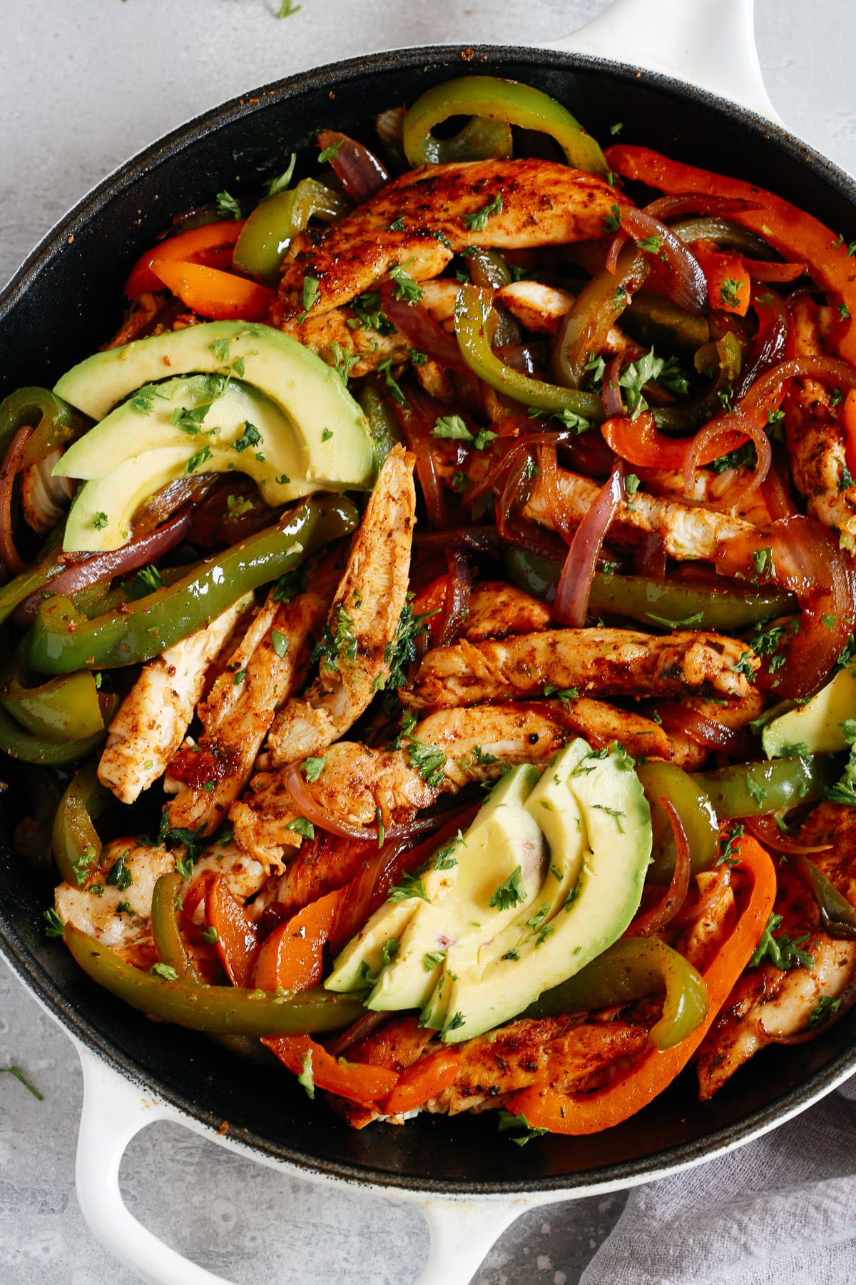 A white skillet with chicken fajitas, bell peppers, and avocado slices.