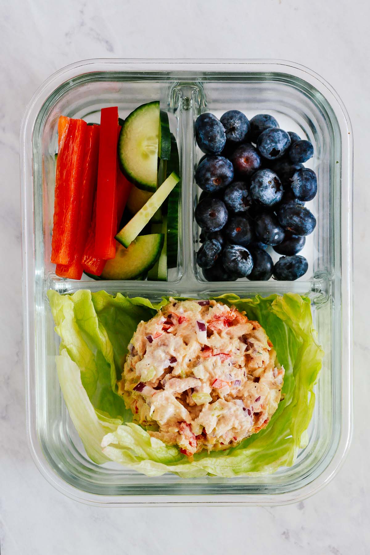 A glass meal prep container with a scoop of tuna salad, blueberries, red peppers, and cucumbers.