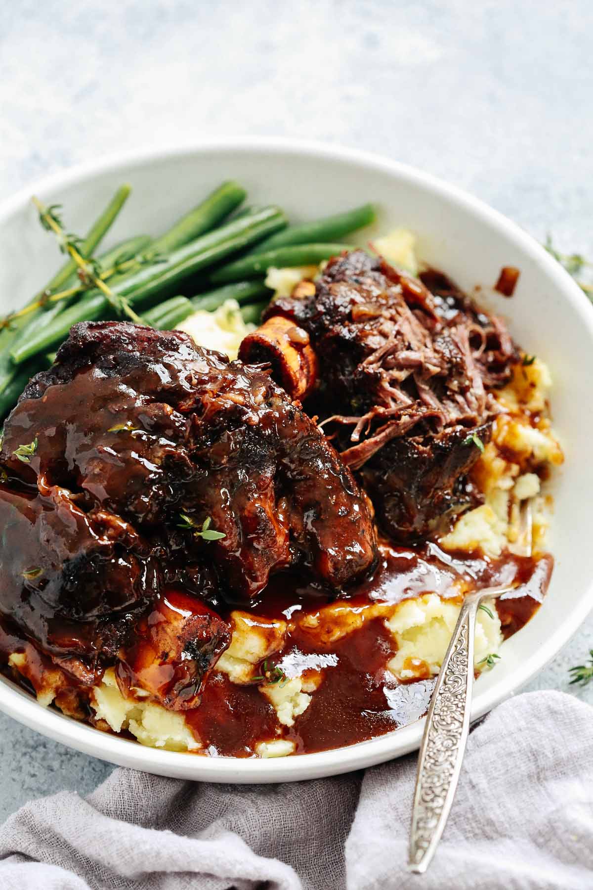 A close up of short ribs with green beans and mashed potatoes in a white bowl