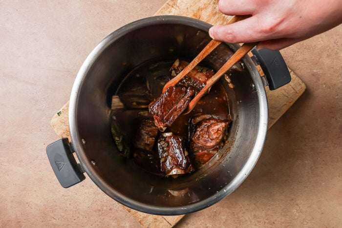 A hand uses tongs to lift a piece of cooked meat from a pot filled with Instant Pot Short Ribs in a rich dark sauce, set on a wooden cutting board atop a brown countertop.