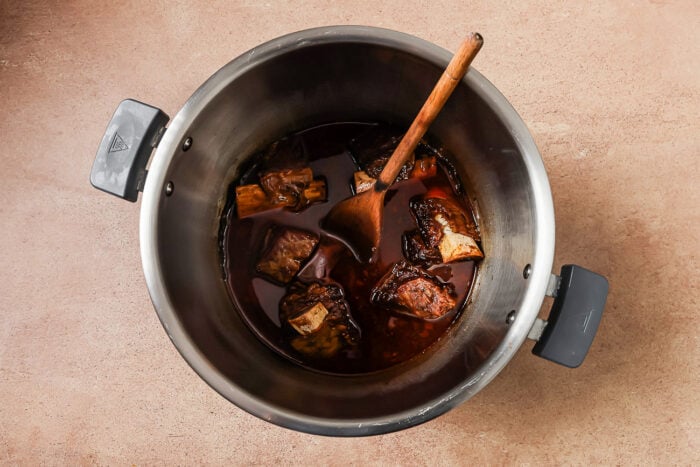 A large metal pot filled with Instant Pot Short Ribs braised in dark broth, with a wooden spoon resting inside, placed on a light brown surface.