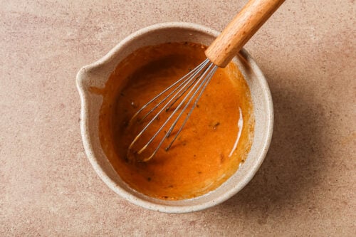 A ceramic bowl containing a reddish-brown sauce or dressing for Instant Pot Short Ribs is being mixed with a metal whisk with a wooden handle, set on a beige countertop.