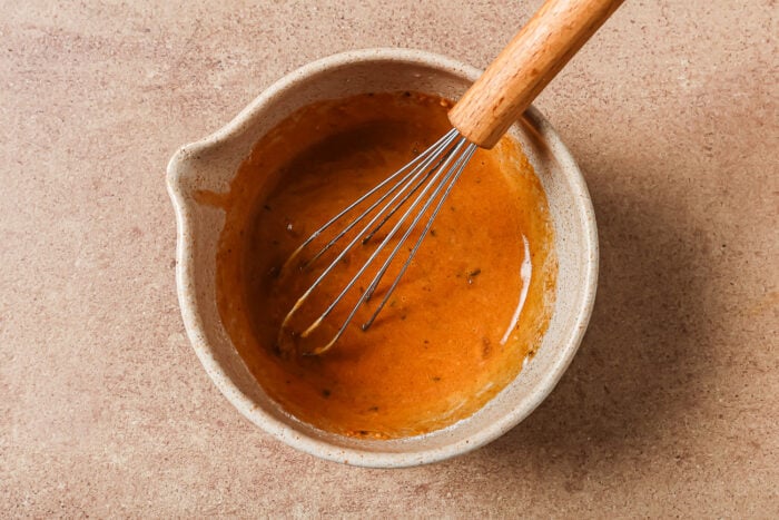 A ceramic bowl containing a reddish-brown sauce or dressing for Instant Pot Short Ribs is being mixed with a metal whisk with a wooden handle, set on a beige countertop.
