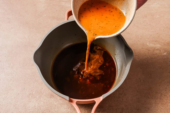 A hand pours an orange-colored sauce from a bowl into a pot containing dark liquid, preparing flavorful Instant Pot Short Ribs on a light brown surface.