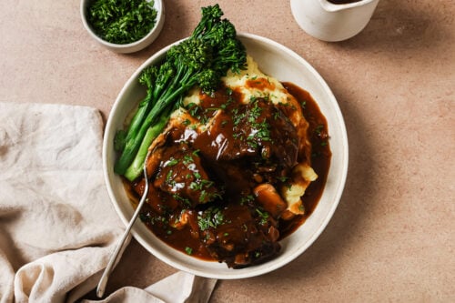 A bowl of mashed potatoes topped with tender Instant Pot Short Ribs in brown gravy, garnished with chopped parsley, served with steamed broccolini. A small bowl of parsley and a beige napkin are nearby.