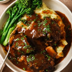 A bowl of mashed potatoes topped with tender Instant Pot Short Ribs in rich brown gravy, garnished with chopped herbs, served with steamed broccolini on the side and a fork resting in the dish.