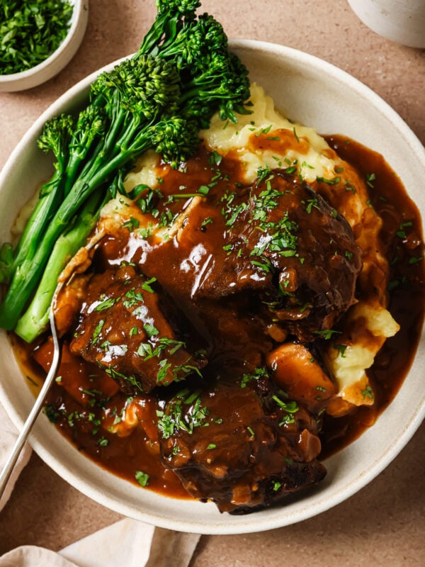 A bowl of mashed potatoes topped with tender Instant Pot Short Ribs in rich brown gravy, garnished with chopped herbs, served with steamed broccolini on the side and a fork resting in the dish.
