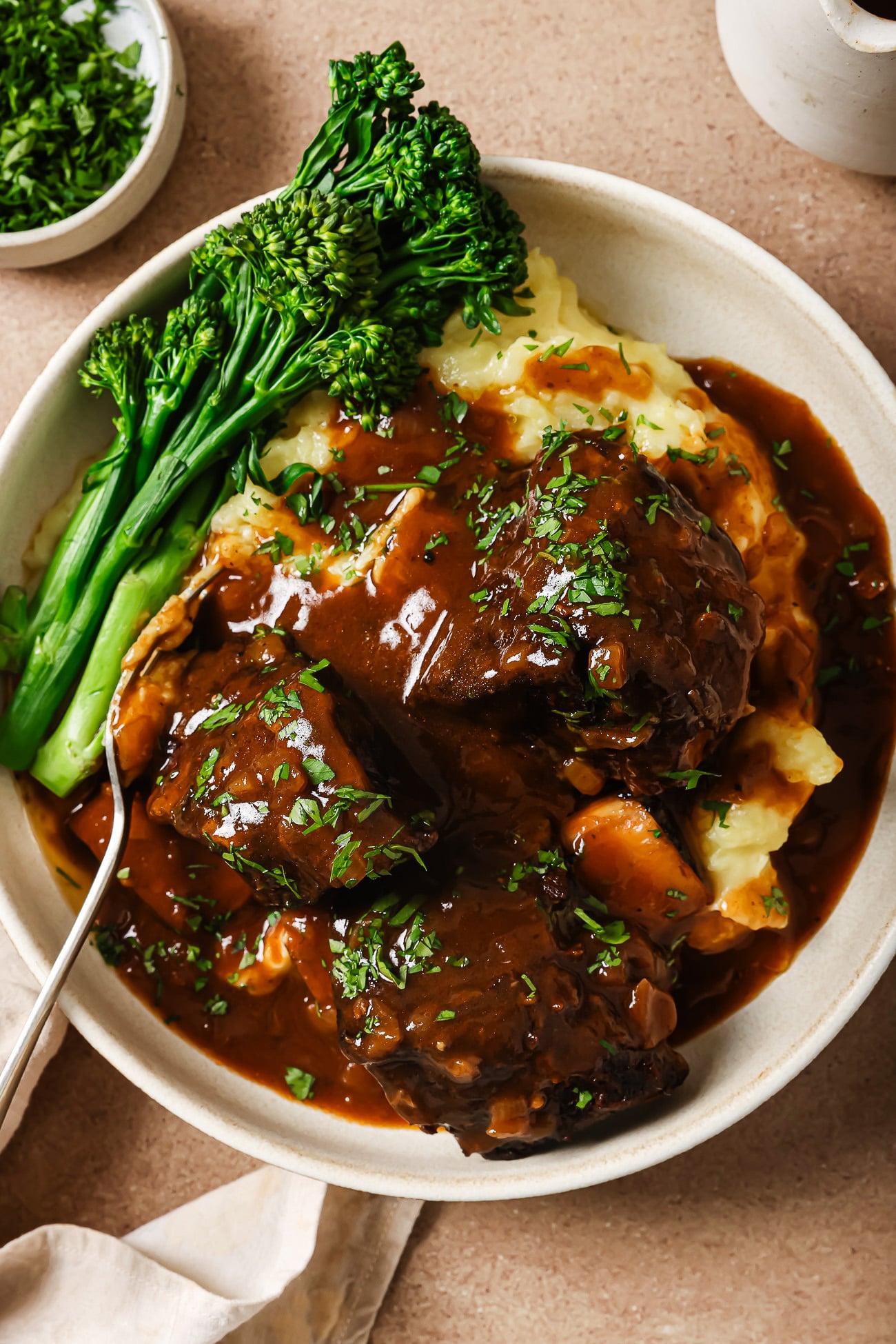 A bowl of mashed potatoes topped with tender Instant Pot Short Ribs in rich brown gravy, garnished with chopped herbs, served with steamed broccolini on the side and a fork resting in the dish.