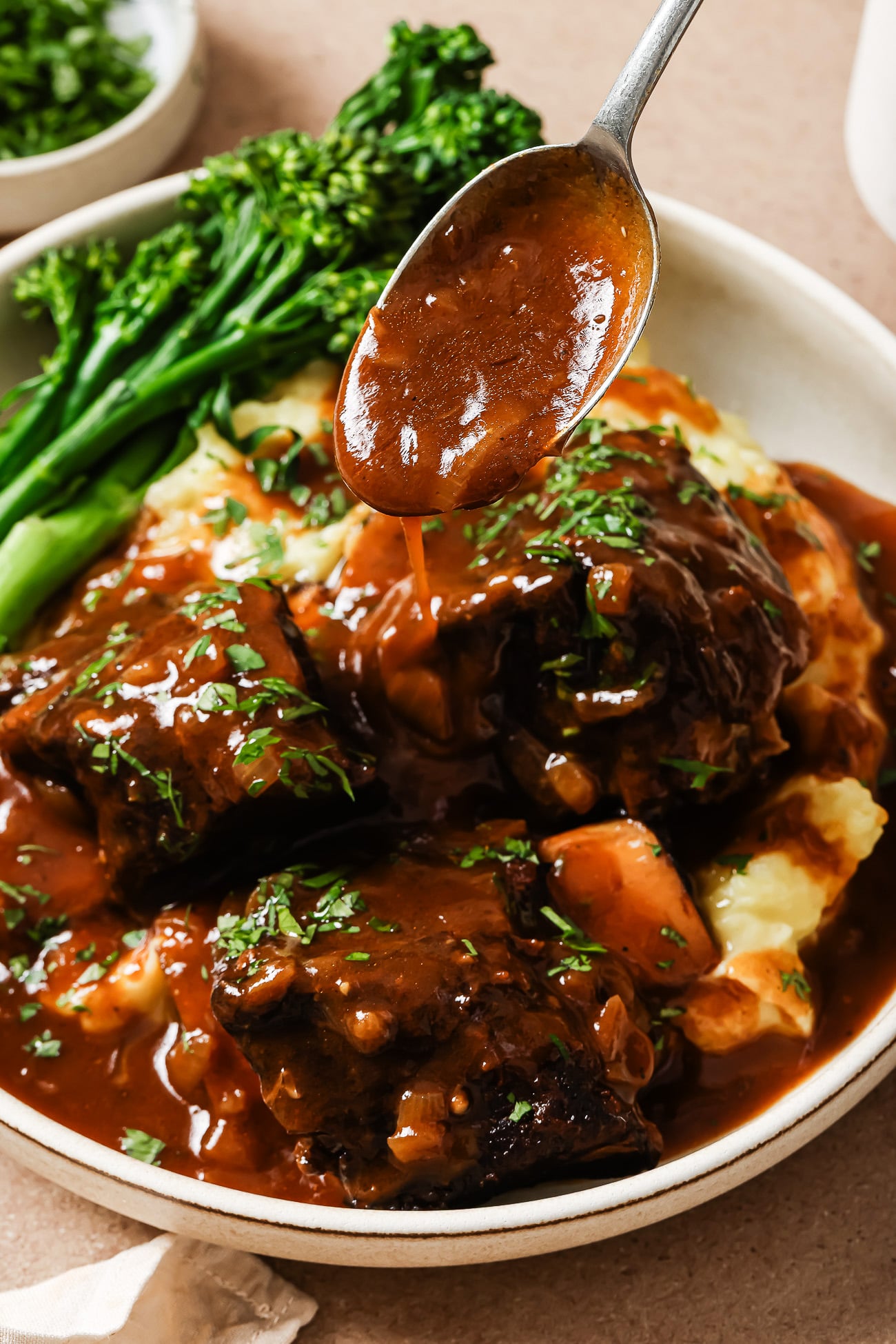 A plate of Instant Pot Short Ribs served over mashed potatoes with gravy being spooned on top, garnished with chopped herbs and accompanied by steamed broccolini.