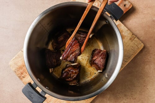 A hand uses wooden tongs to lift a browned piece of Instant Pot Short Ribs from a pot containing several other browned short ribs on a wooden cutting board.