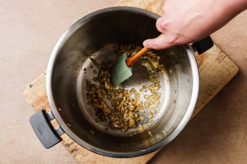 A hand stirs sautéed chopped onions in a large stainless steel pot, preparing the base for Instant Pot Short Ribs. The pot rests on a wooden cutting board placed on a beige countertop.