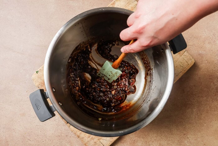 A hand stirs a dark, caramelized mixture of onions in a stainless steel pot with a spatula, prepping the base for savory Instant Pot Short Ribs on a wooden cutting board atop a brown countertop.
