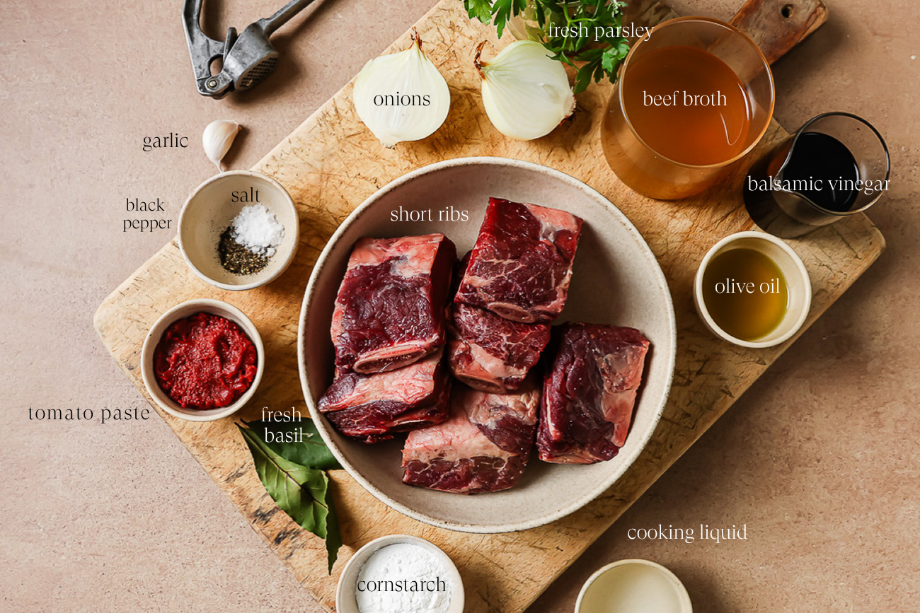 A bowl of raw Instant Pot Short Ribs sits on a wooden board, surrounded by labeled ingredients: tomato paste, fresh basil, black pepper, salt, garlic, cornstarch, onions, beef broth, balsamic vinegar, olive oil, cooking liquid, and fresh parsley.