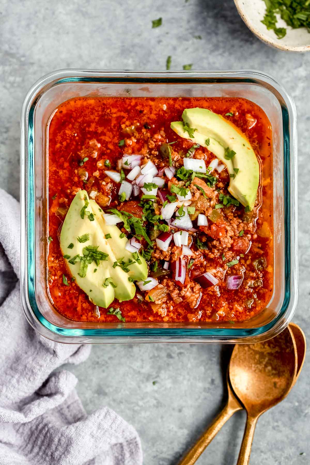 glass container holding one serving of chili with avocado slices and onions