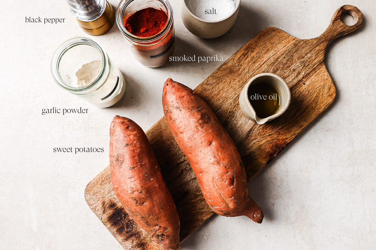 Two whole sweet potatoes on a wooden cutting board, ready to become delicious roasted sweet potato, with jars of black pepper, smoked paprika, garlic powder, salt, and a small container of olive oil arranged nearby on a light surface.