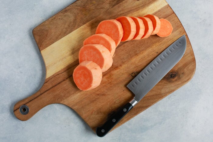 A wooden cutting board with evenly sliced roasted sweet potato pieces and a chef’s knife resting beside them on a light gray surface.