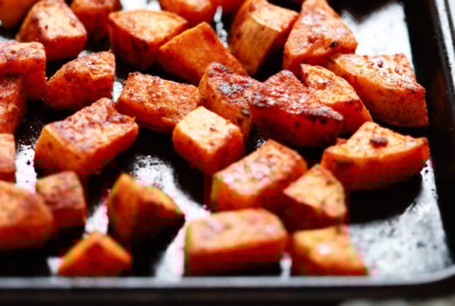 Close-up of roasted sweet potato cubes seasoned with spices on a dark baking sheet, highlighting the golden roasted sweet potato in sharp focus upfront and a blurred background.