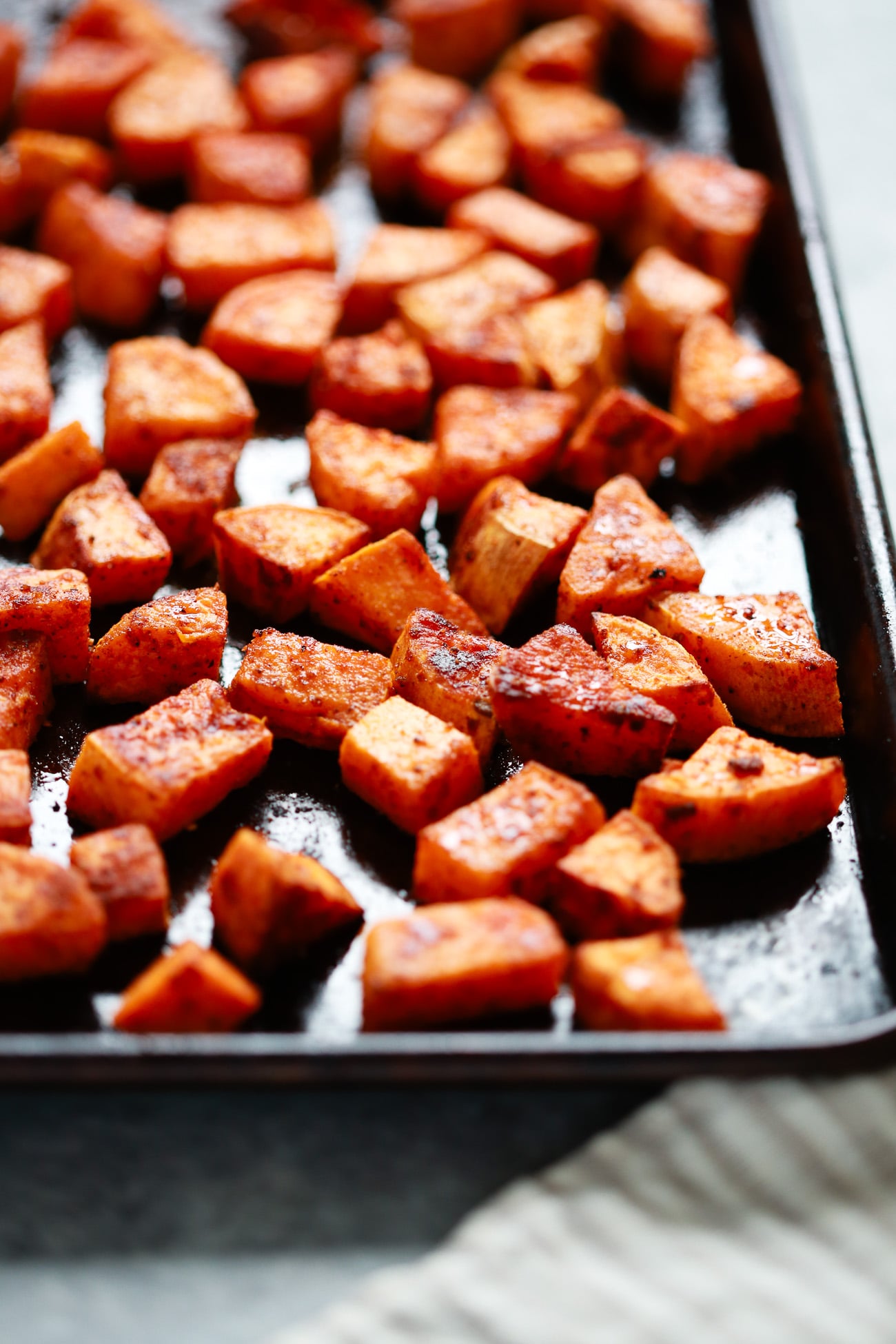 Close-up of roasted sweet potato cubes seasoned with spices on a dark baking sheet, highlighting the golden roasted sweet potato in sharp focus upfront and a blurred background.
