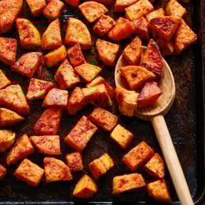 Chunks of roasted sweet potato on a dark baking sheet, some scooped up with a wooden spoon. The potatoes are golden brown and seasoned, with a striped kitchen towel partially visible in the corner.