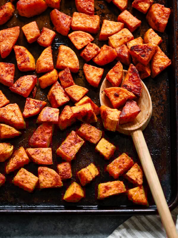 Chunks of roasted sweet potato on a dark baking sheet, some scooped up with a wooden spoon. The potatoes are golden brown and seasoned, with a striped kitchen towel partially visible in the corner.
