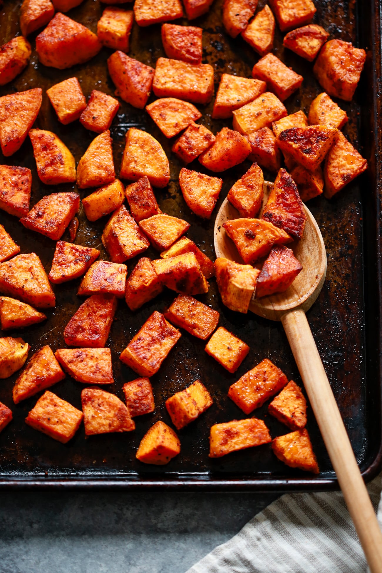 Chunks of roasted sweet potato on a dark baking sheet, some scooped up with a wooden spoon. The potatoes are golden brown and seasoned, with a striped kitchen towel partially visible in the corner.
