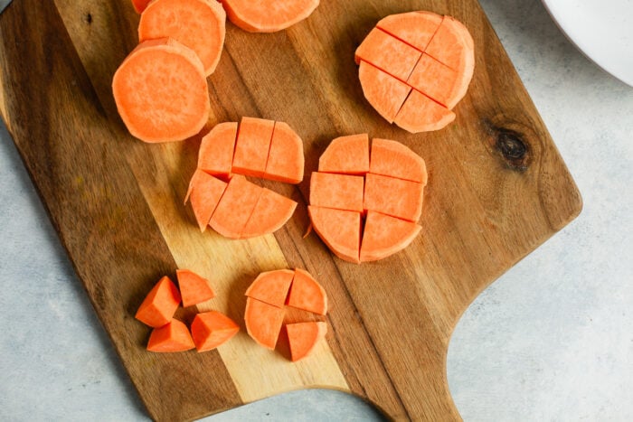 Sliced sweet potatoes on a wooden cutting board, some pieces cut into cubes and others into circles, perfect for making roasted sweet potato, with a light gray background.