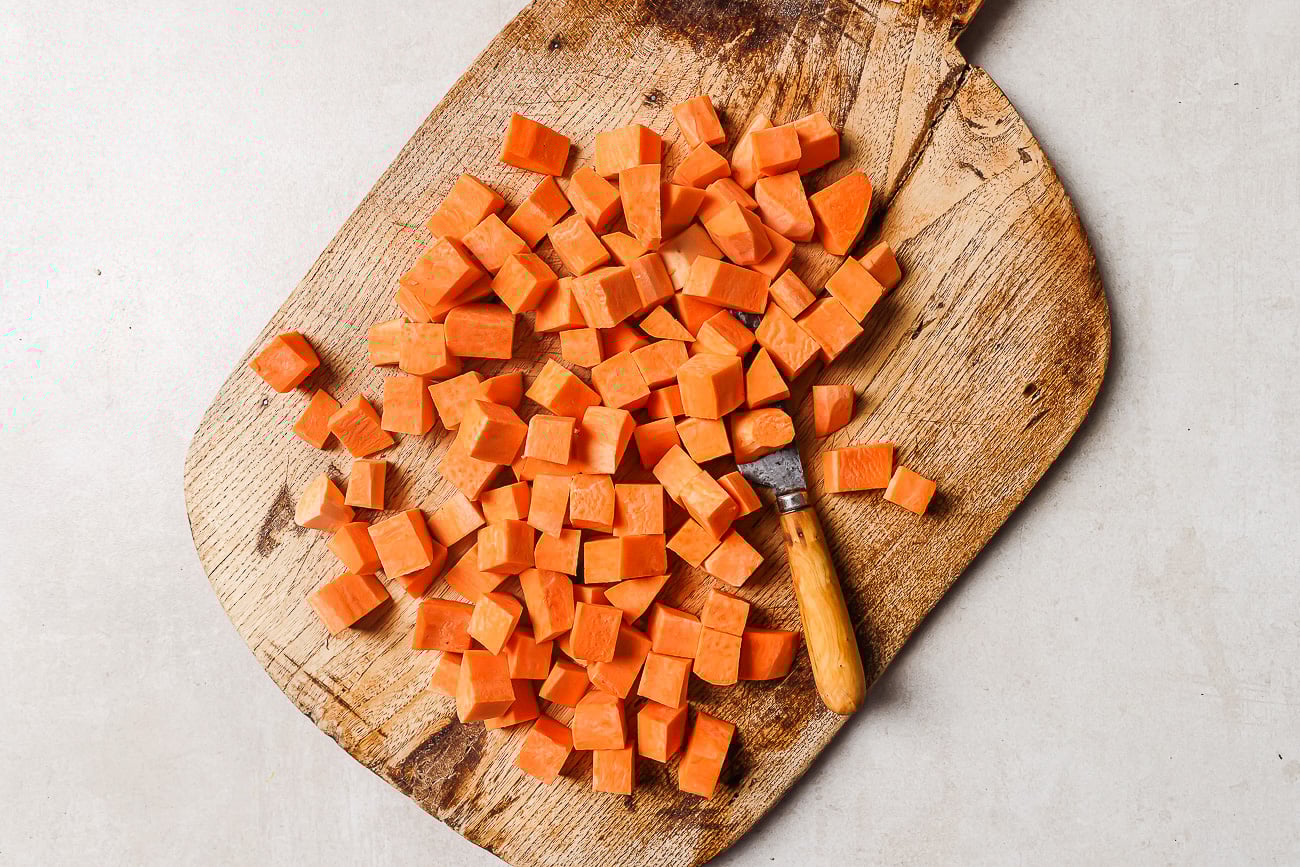Cubed sweet potatoes, perfect for making roasted sweet potato, are spread on a rustic wooden cutting board with a small knife placed among them, all set on a light-colored surface.