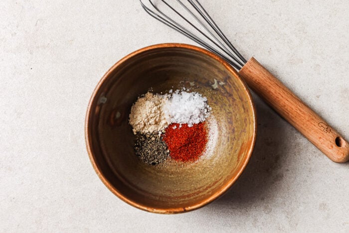 A brown bowl contains four piles of spices—black pepper, white salt, paprika, and garlic powder—perfect for seasoning roasted sweet potato. A metal whisk with a wooden handle rests beside the bowl on a light countertop.