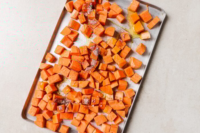 Diced roasted sweet potato pieces are spread on a metal baking sheet, drizzled with olive oil and sprinkled with spices, ready to be baked to perfection.
