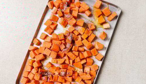 Diced roasted sweet potato pieces are spread on a metal baking sheet, drizzled with olive oil and sprinkled with spices, ready to be baked to perfection.