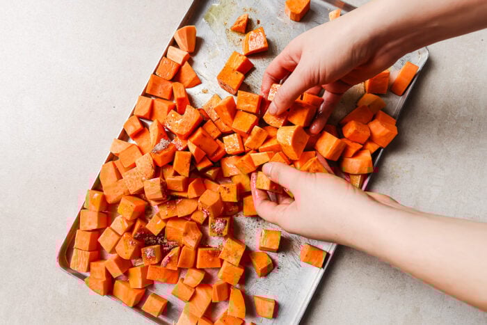 Hands tossing cubed sweet potatoes with seasoning on a metal baking sheet, preparing roasted sweet potato on a light-colored countertop.