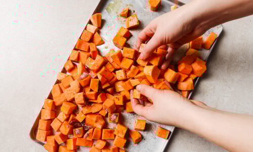 Hands tossing cubed sweet potatoes with seasoning on a metal baking sheet, preparing roasted sweet potato on a light-colored countertop.