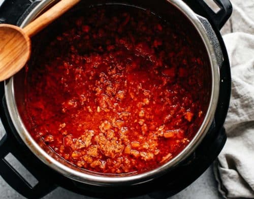 A pot of red chili with ground meat and beans, reminiscent of a hearty Instant Pot Meat Sauce, sits on a stove with a wooden spoon resting on the edge and a beige cloth nearby.