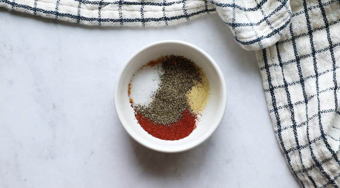 A small white bowl with four different spices—salt, black pepper, paprika, and garlic powder—arranged separately. Perfect for a Roasted Cauliflower Recipe, the bowl sits on a white surface next to a white and blue checkered kitchen towel.
