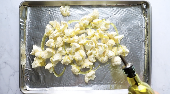 Cauliflower florets on a foil-lined baking sheet being drizzled with olive oil from a bottle, ready for roasting—perfect for any roasted cauliflower recipe.