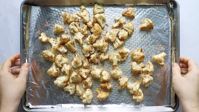 Top-down view of two hands holding a baking sheet lined with foil, filled with roasted cauliflower florets seasoned with spices—a delicious Roasted Cauliflower Recipe ready to enjoy.