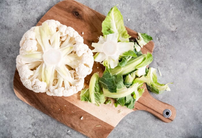 A cauliflower with its stem and leaves partially removed sits on a wooden cutting board, surrounded by discarded leaves and stems, on a gray countertop—ready to be transformed into spicy roasted cauliflower.