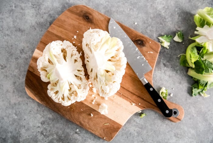 A halved cauliflower and a kitchen knife rest on a wooden cutting board, with a few cauliflower leaves and crumbs scattered on a gray countertop—perfectly prepped for making Spicy Roasted Cauliflower.