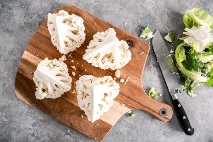 Four Spicy Roasted Cauliflower wedges on a wooden cutting board, with a knife and cauliflower leaves beside them on a gray surface.