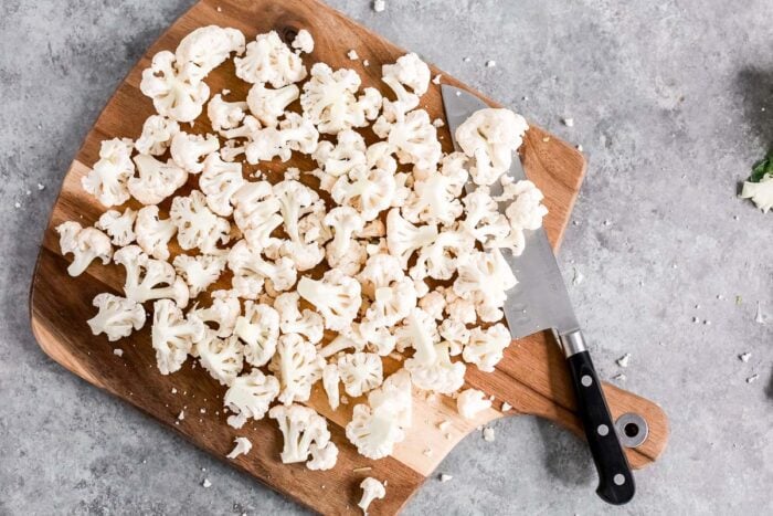 Chopped cauliflower florets, ready for a Spicy Roasted Cauliflower dish, are spread on a wooden cutting board with a chef’s knife beside them, all set on a gray countertop.
