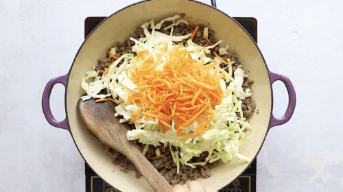 Shredded cabbage and carrots added on top of cooked ground beef in a skillet.