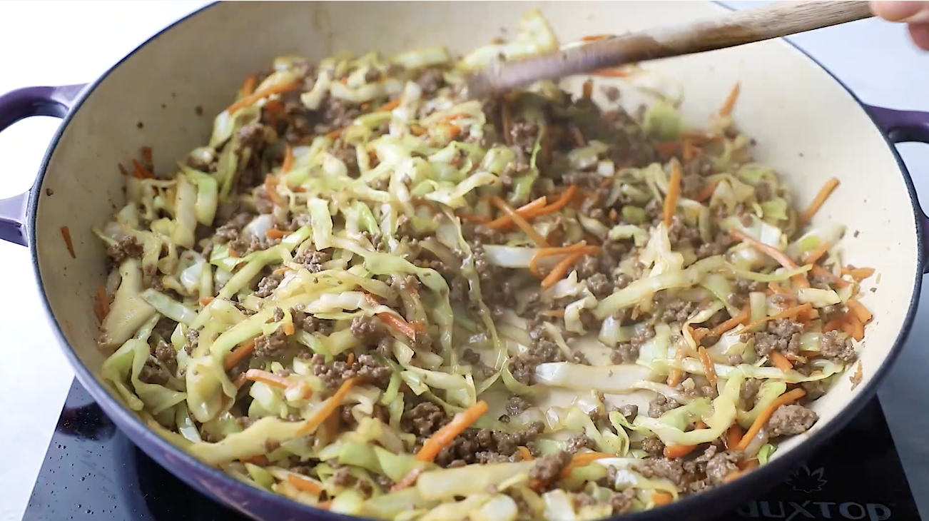 Cooking ground beef in a skillet with a wooden spoon as the first step for making Egg Roll in a Bowl.