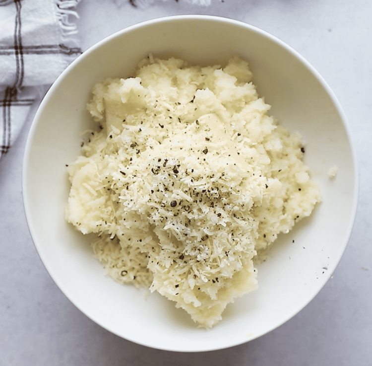 overhead view of a white bowl containing mashed cauliflower, parmesan and spices