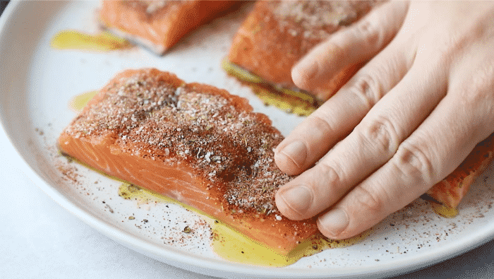 A hand pressing a dry rub into a raw salmon fillet on a white plate to prepare a blackened salmon recipe.