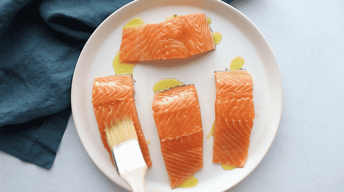 Four salmon fillets for a blackened salmon recipe on a white plate being brushed with oil.