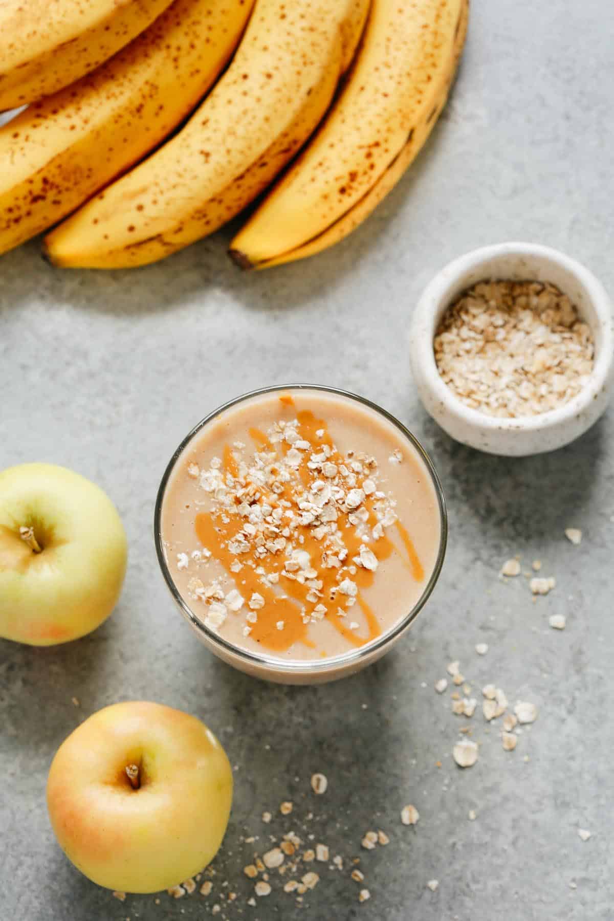 cup of apple smoothie with oats sprinkled on top with bananas, apples, and oats placed beside it