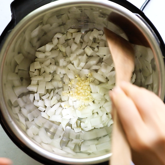 Chopped onions and minced garlic are in an Instant Pot, with a hand stirring them using a wooden spatula.