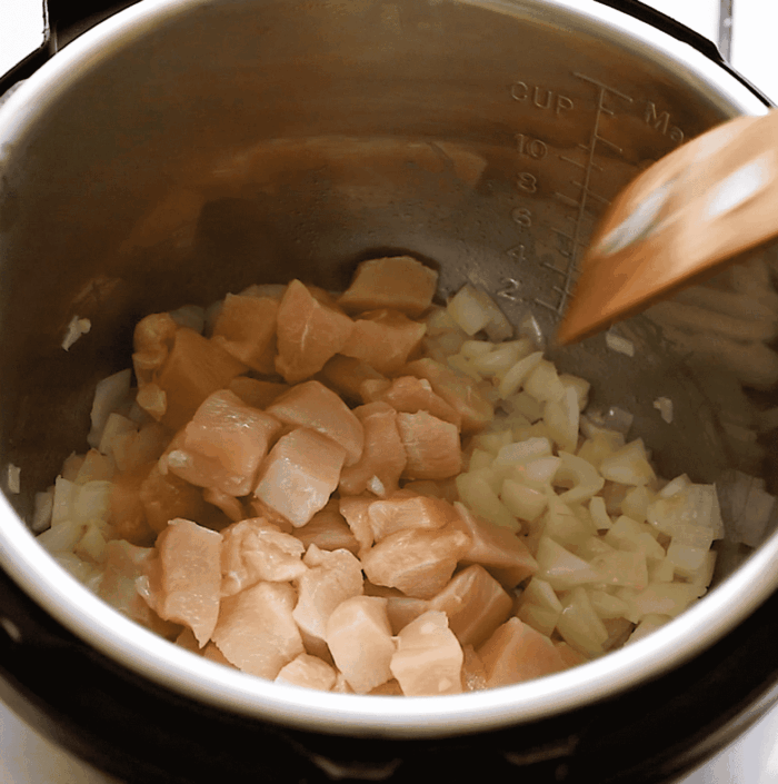 Diced raw chicken and chopped onions are being stirred with a wooden spatula inside a metal pot, likely an Instant Pot or pressure cooker.